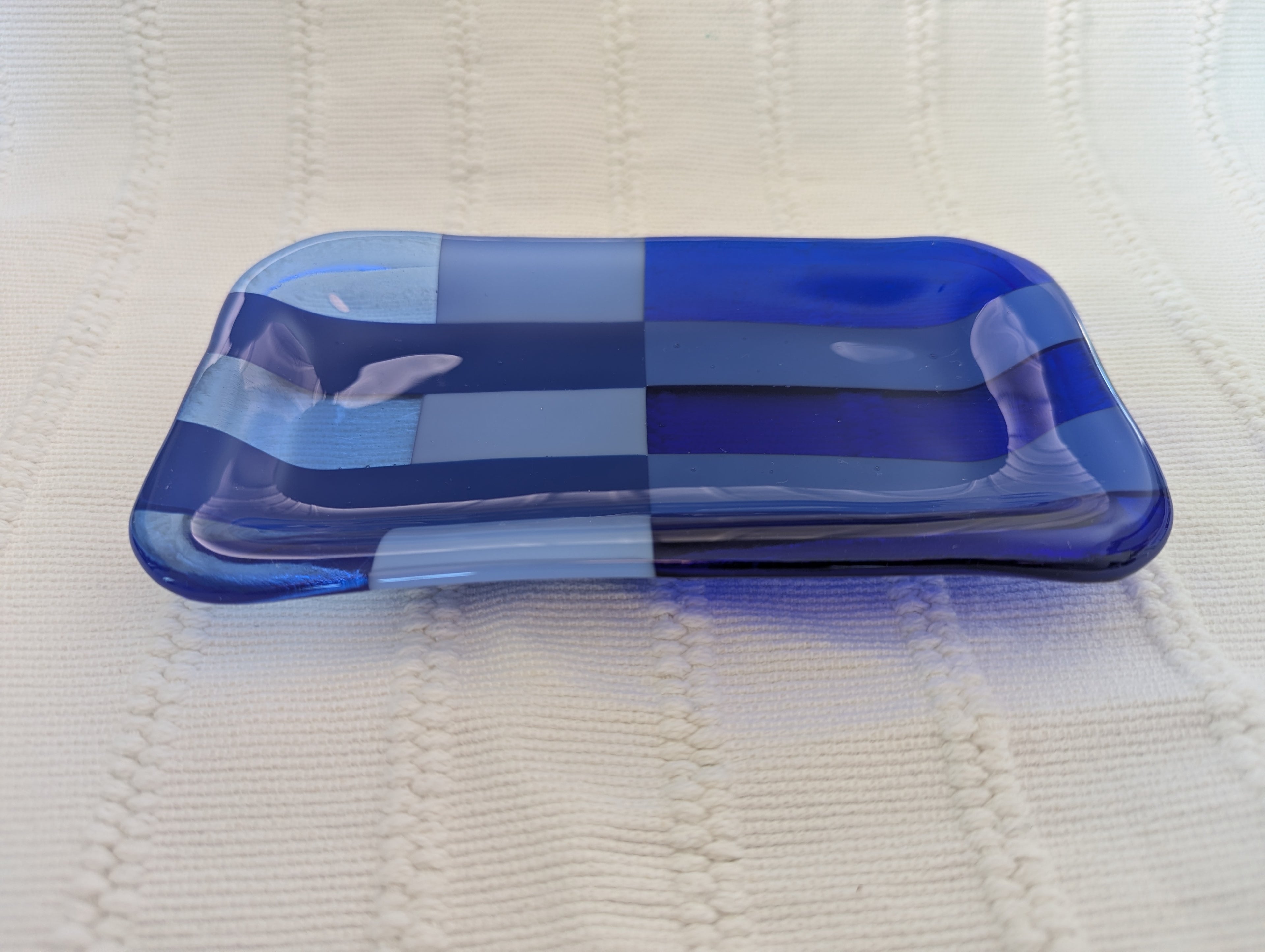 A rectangular glass tray with rounded corners, featuring a patchwork design of various shades of blue and light blue translucent glass panels, resting on a textured cream-colored knit blanket.