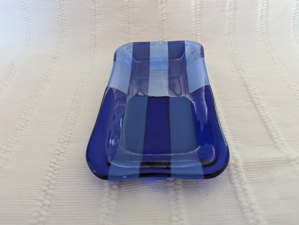A rectangular glass dish with rounded corners, featuring alternating vertical stripes of translucent light blue, opaque medium blue, and deep cobalt blue, resting on a textured cream-colored knit blanket.