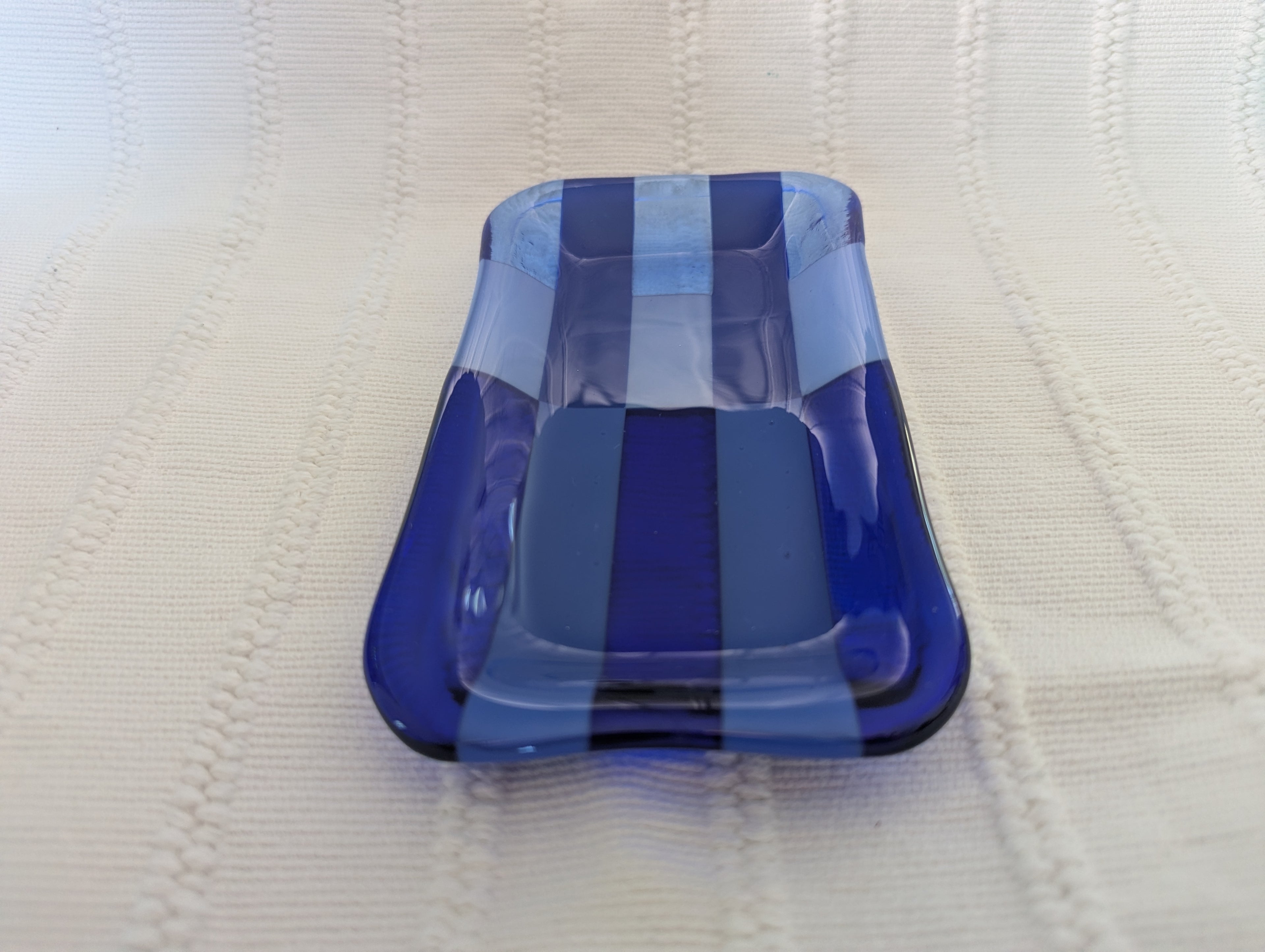 A rectangular glass dish with rounded corners, featuring alternating vertical stripes of translucent light blue, opaque medium blue, and deep cobalt blue, resting on a textured cream-colored knit blanket.
