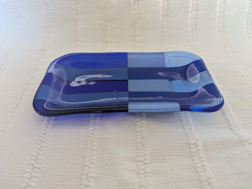 A small rectangular glass dish with rounded corners in a blue and white checkerboard pattern, resting on a textured cream-colored knit blanket.