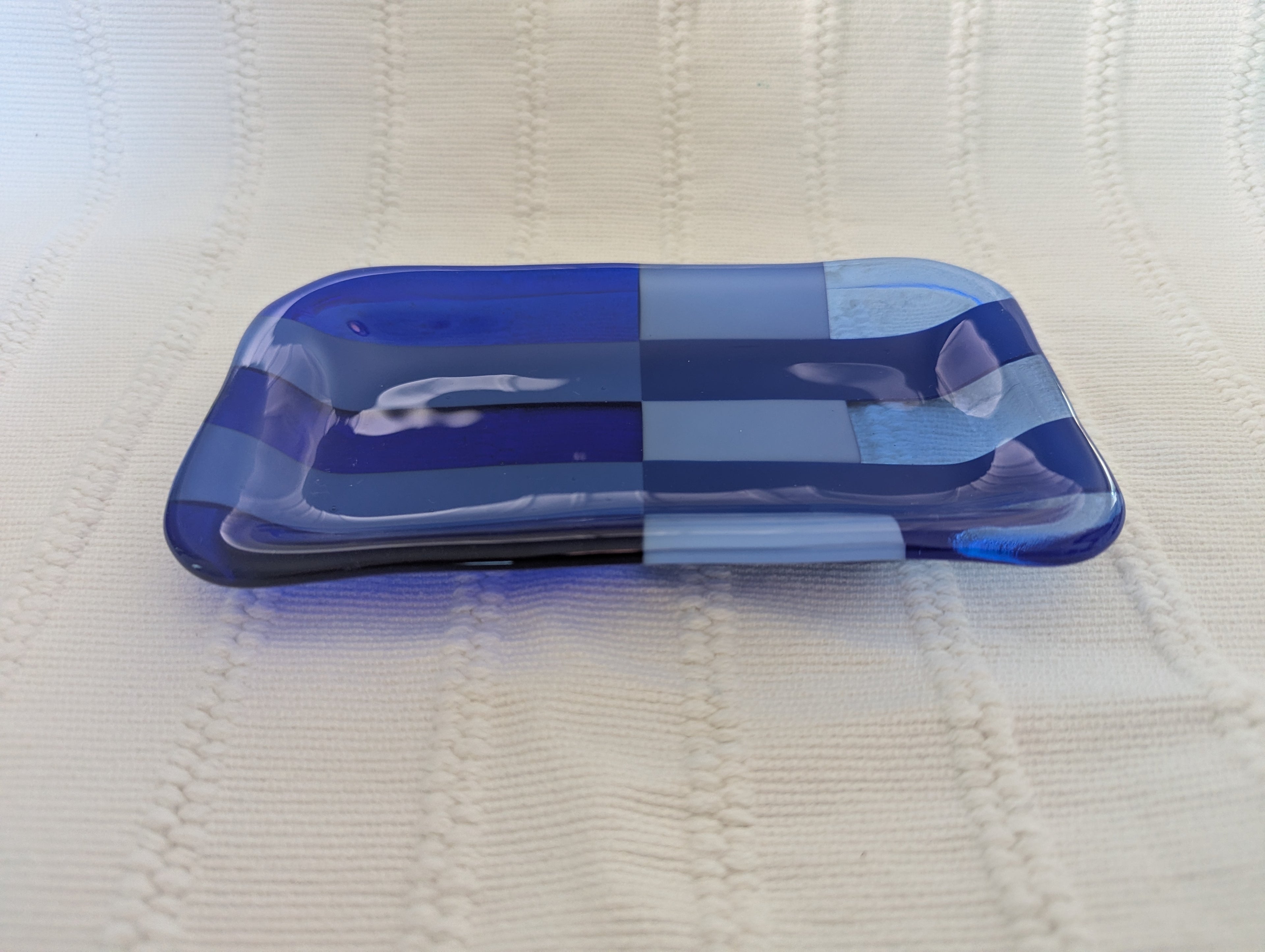 A small rectangular glass dish with rounded corners in a blue and white checkerboard pattern, resting on a textured cream-colored knit blanket.
