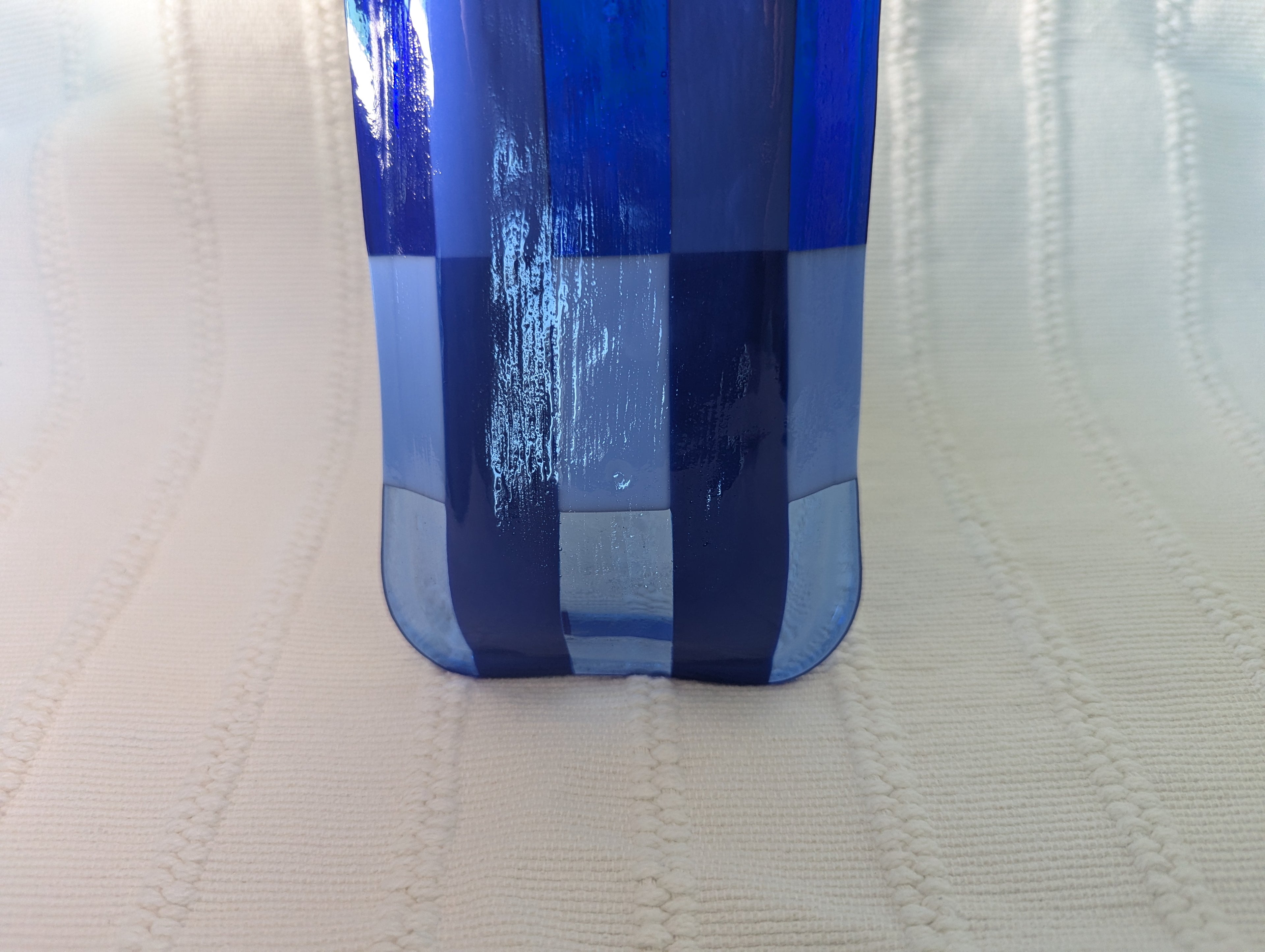 A close-up of a blue and light blue checkered glass object standing on a textured cream-colored surface.