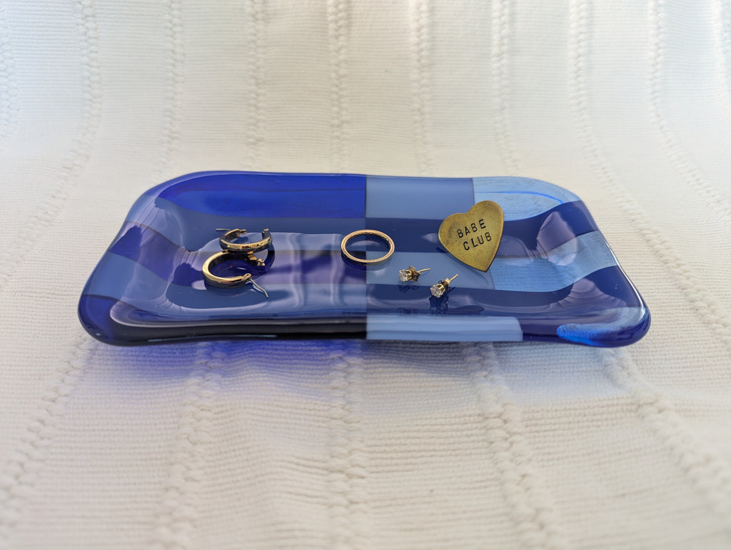 A blue translucent glass jewelry tray with a heart-shaped piece engraved with 'BASE CLUB' resting on a textured cream knit blanket, alongside several gold rings and earrings.