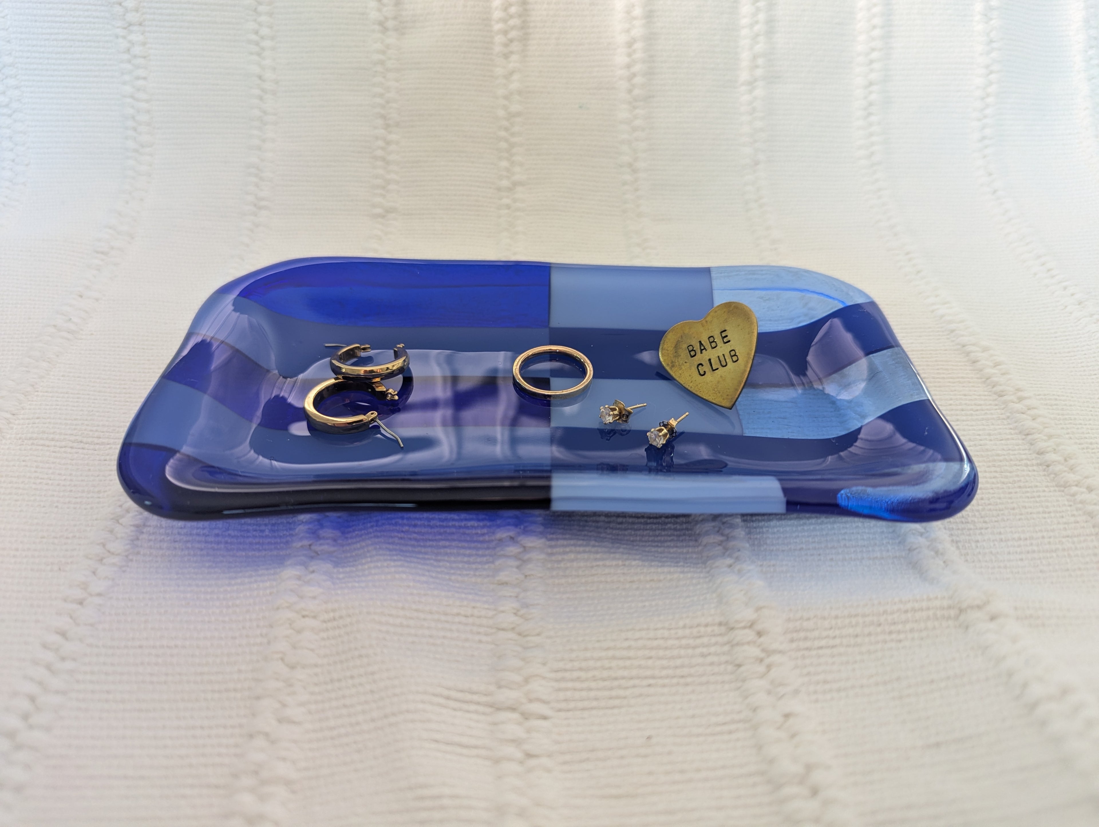 A blue translucent glass jewelry tray with a heart-shaped piece engraved with 'BASE CLUB' resting on a textured cream knit blanket, alongside several gold rings and earrings.