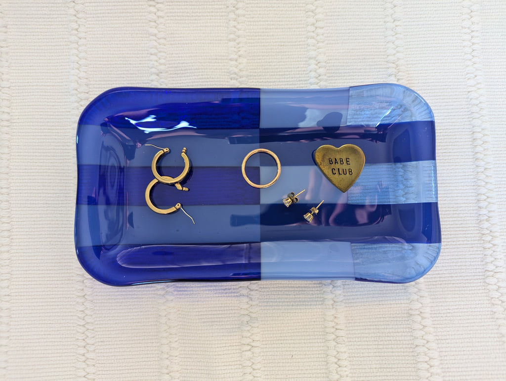 A set of gold-toned jewelry pieces, including hoop earrings, a ring, and a heart-shaped charm engraved with 'BARE CLUB', displayed on a blue striped glass tray over a white textured blanket.