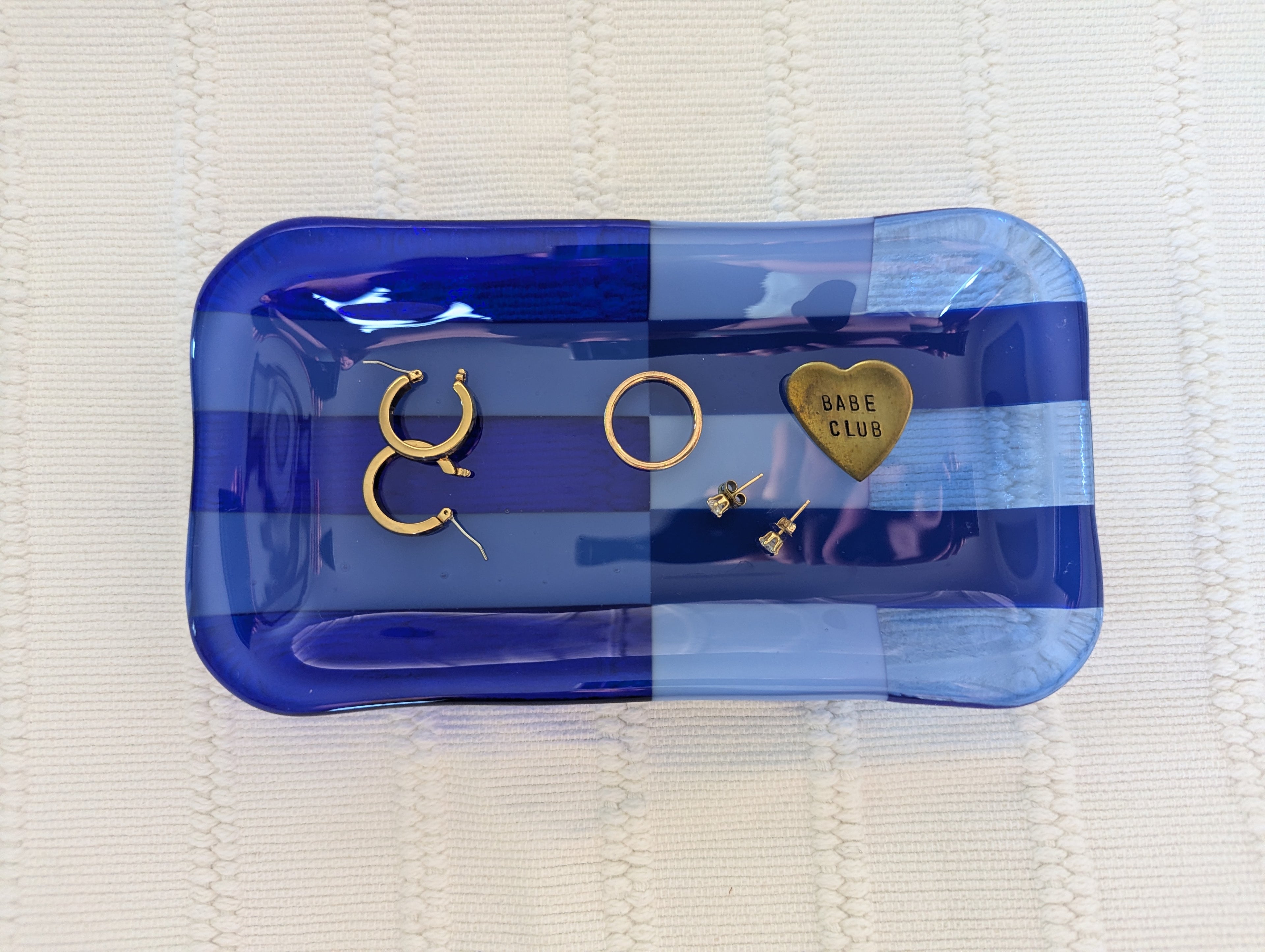 A set of gold-toned jewelry pieces, including hoop earrings, a ring, and a heart-shaped charm engraved with 'BARE CLUB', displayed on a blue striped glass tray over a white textured blanket.